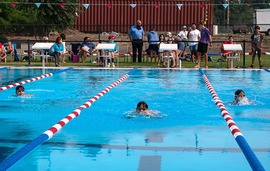 Three children race in lanes in a swimming pool while people on land watch