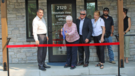 Five people watch as another person uses oversized scissors to cut a red ribbon stretched across the entrance to the cemetery office