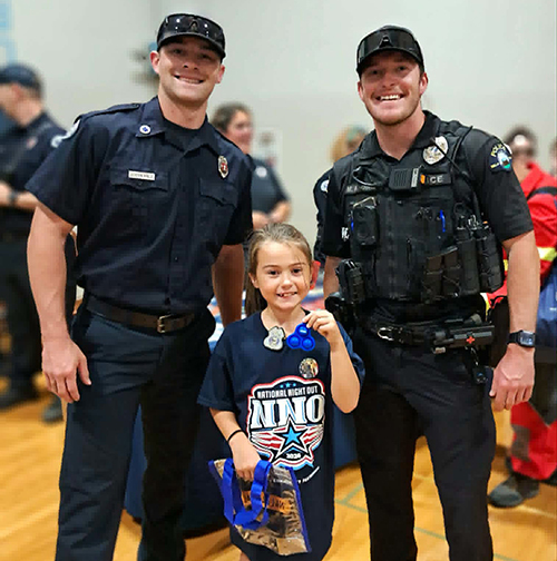 A child wearing a National Night Out T-shirt poses for a photo with a firefighter and a police officer