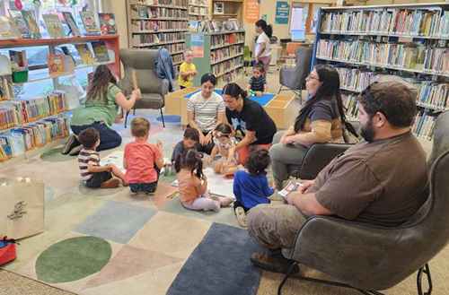 Young children sit on the floor of the temporary library with parents around them during Bilingual Storytime