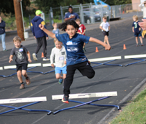 A preadolescent child leaps over a hurdle on a track while two other kids run behind