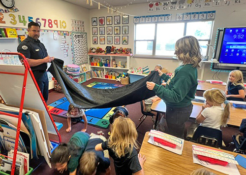 A firefighter and a teacher hold a blanket horizontally between them while children crawl beneath it to teach smoke safety