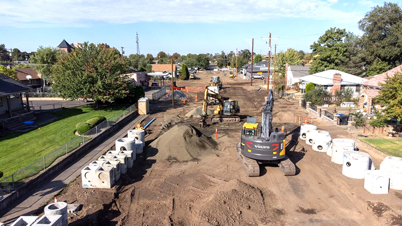 Crews with heavy equipment work to install water main in a gravel-surfaced roadway