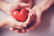 Two pairs of hands holding a wooden carved heart that is painted red.