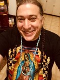 Brian Frisina, smiling, with a long braid, wears a shell necklace, a black t-shirt with artwork of the Holy Mother Mary printed on the front.