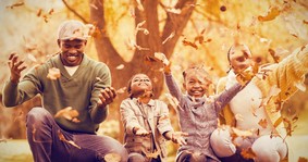 Photo of a family smiling and playing in the autumn leaves in a sunny park.