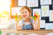 A child holds her hands up, covered in colorful paint, surrounded in artwork on the walls and table. 
