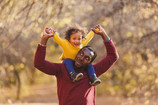 A father happily looks up at his toddler sitting on his shoulders, as they hold clasped hands in an excited 'V' above their heads.