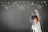 Photo of a little girl standing in front of a chalkboard with an astronaut suit, stars and space-themed doodles drawn around her.