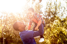 Photo of a father holding up his smiling toddler above his head, backlit by a bright, sunny day.