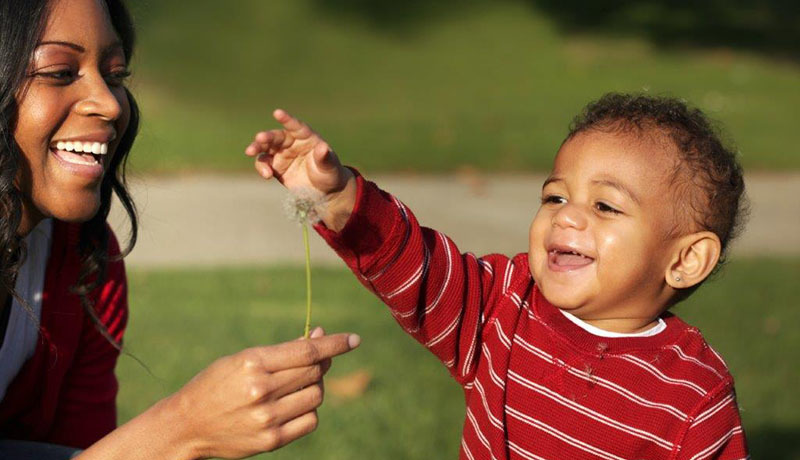 A mother holds up a dandelion to her smiling toddler. 