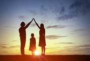 Photo of two parents, facing each other with hands clasped high above a small child, stand silhouetted against a bright orange sunset. 