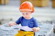 Photo of a toddler wearing an orange hard hat, looking at a paper in deep concentration.