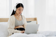 Photo of woman wearing headphones on laptop, sitting up in bed