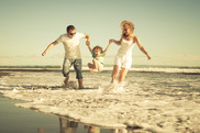 Photo of two parents holding the hands of their child, as they run across the beach shore, holding their child playfully above the waves.