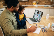 Photo of a father and toddler looking at a laptop together, interacting with a health care provider over video chat.