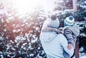 A parent and child bundled up in winter gear look out at a sunlit, snowy nature trail scene.