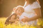 A little girl leans against her mother's pregnant belly, as they sit together in a sunlit field.