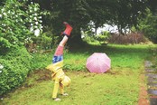 A child in a yellow raincoat and red rainboots does a handstand in a green, grassy yard, their umbrella open and visible in the background.