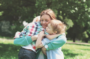 A smiling mother hugs her two young children as they sit in a sunny park.