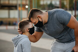 Photo of a father leaning down, forehead to forehead with his son, both wear black protective kn95 face masks.