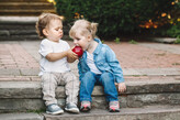 Photo of a toddler holding a red apple toward another toddler’s open mouth.