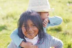 Photo of two Native American Indian children in a sunlit field. The child in front smiles at the camera, the other playfully hides behind.