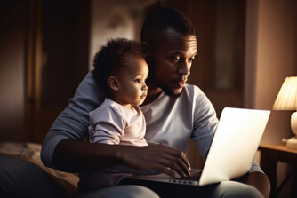 Photo of a baby sitting on their father's lap, both looking intently at a laptop, in a softly lamp-lit room.