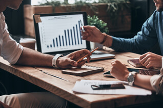 Coworkers surrounding a table, discussing a data chart displayed on a laptop. Medium shot, cropping off their heads, focusing on their hands.