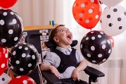 Young boy in wheelchair laughs and smiles looking up, surrounded by red, black and white polka dot balloons.