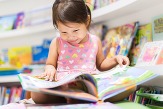 Toddler girl looks intently at a big, colorfully illustrated picture book in her lap.