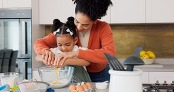 Mother and daughter cracking eggs together into a bowl, at a kitchen counter, surrounded in baking supplies and ingredients. 