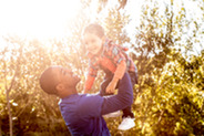 A father holds his smiling toddler up toward the sky, surrounded in a sunny, nature setting.