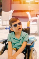 Young boy in wheelchair wears sunglasses in a bright, sunny room.