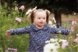 A toddler girl in pigtails and blue floral top, happily stands in a flower field with her arms outstretched.