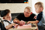 A father happily looks at his two young sons playing, one sits in a wheelchair and the other stands, all around a small table.