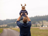 A father stands with his back to the camera, his arms raised in mid-toss-and-catch, as his toddler hangs mid-air above him, pigtails flying. 