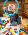 A toddler girl in blonde pigtails, sits on the floor, holding up a turquoise hoop and surrounded by colorful blocks and toys.