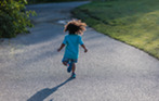 A little boy, with dark curly, shoulder-length hair, a turquoise t-shirt and blue jean shorts, happily runs away from the camera and down a sun