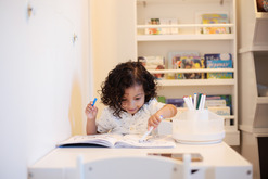 A little boy with dark, curly, shoulder-length hair and turquoise t-shirt concentrates while happily coloring with markers in a coloring book.