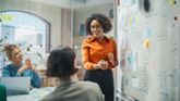 Three coworkers meet and discuss around a table, by a big whiteboard covered in notes.