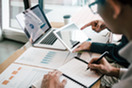 Coworkers surrounding a table, discussing a data chart displayed on a laptop. Medium shot, cropping off their heads, focusing on their hands.