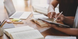 Picture of books, paper, sticky notes covering desk as two people take notes and work on computer.