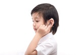 A young boy in profile, wearing a crisp, white polo shirt, reaches up to adjusting his hearing aid.