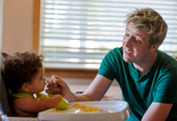 A father smiles, sitting next to a highchair and holding a small piece of food, encouraging his curious toddler to eat.