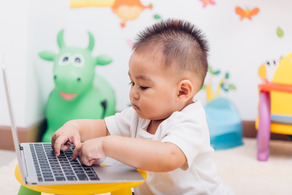 Chubby-cheeked Asian baby sitting in a playroom, in deep concentration, looking down at the keyboard of a computer laptop.