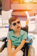A young boy in a wheelchair, wearing sunglasses, sitting on a sunlit patio.