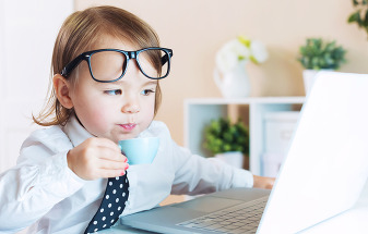 Toddler wearing a white button up shirt, glasses and navy blue and white polka dot tie, sips from a tiny teacup, looking intently at an open laptop.
