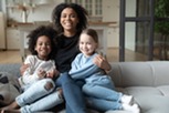Portrait of black mom hugging two little daughters, sitting on cozy couch in living room, looking at camera, multiracial family