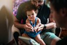 Two fathers read to their baby, who is looking curiously and intently at the book.